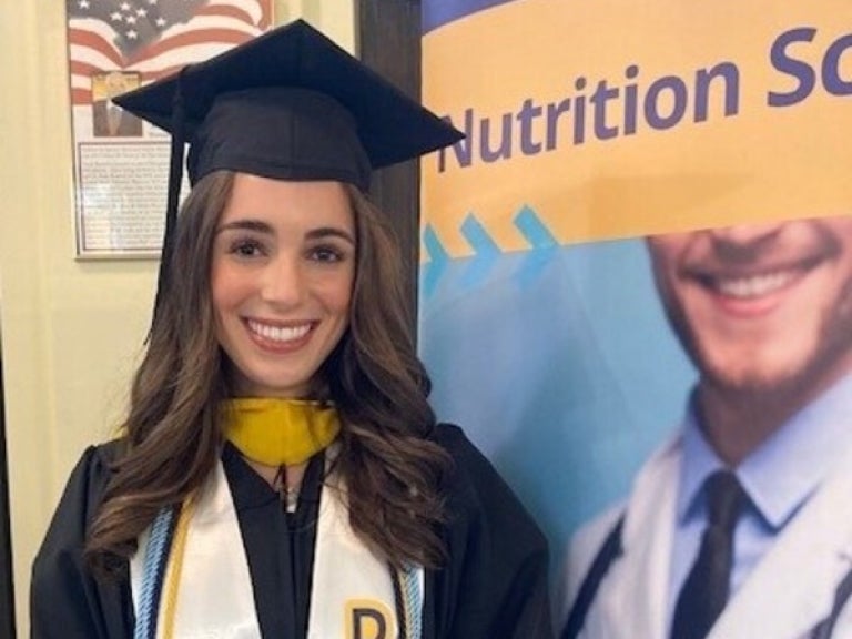 Giavanna Gulino in her graduation cap and gown next to a nutrition science sign.