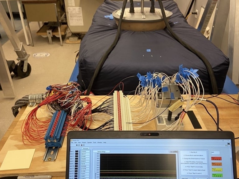Equipment set up for the hemispherical indenter test. The hemispherical indenter is resting on a mattress while a laptop runs a LabVIEW program that is graphing pressure data from each sensor.