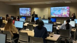 Students sitting in a computer lab with desktop computers, listening to someone give a lecture.