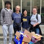Three students and a faculty member standing behind a cart full of food boxes posing for a group photo.