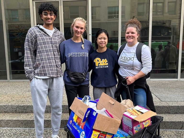 Three students and a faculty member standing behind a cart full of food boxes posing for a group photo.