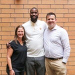 Three people posing for a group photo in front of an orange-brick wall.