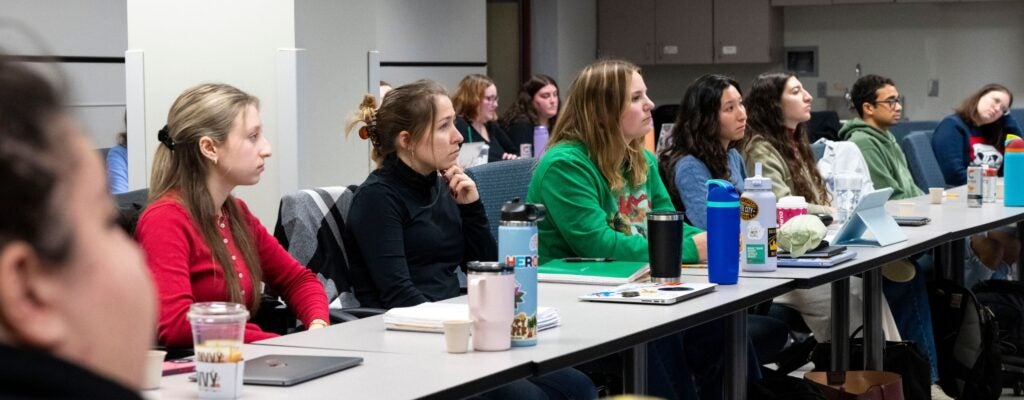 Students sitting next to one another at a long lecture table listening to a speaker not visible in the image.
