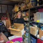 Woman with blonde hair and glasses holding a box of décor in a full storage unit.