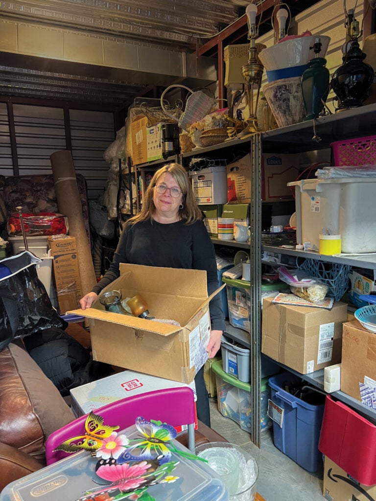 Woman with blonde hair and glasses holding a box of décor in a full storage unit.