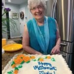 A woman with gray hair and glasses stands in front of a cake