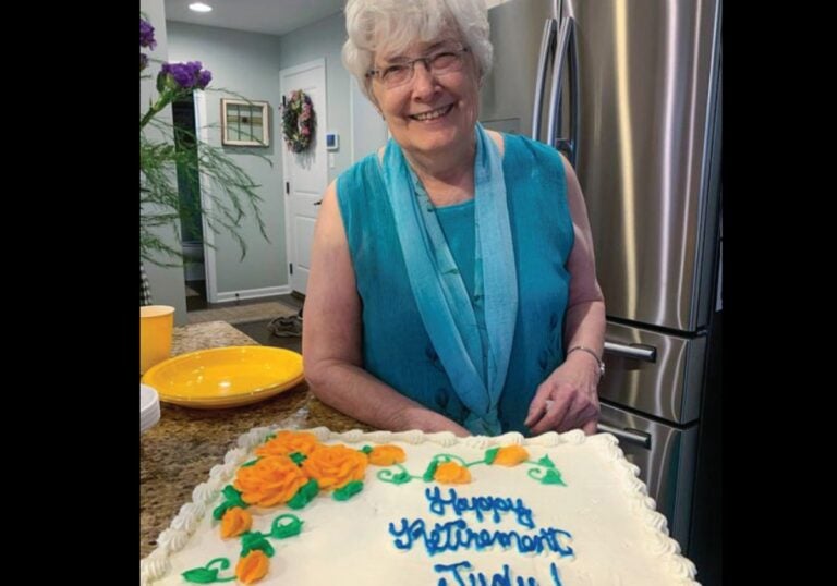 A woman with gray hair and glasses stands in front of a cake