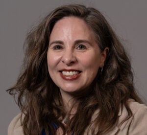 Woman with wavy, long brown hair wearing a brown blazer and royal blue blouse.