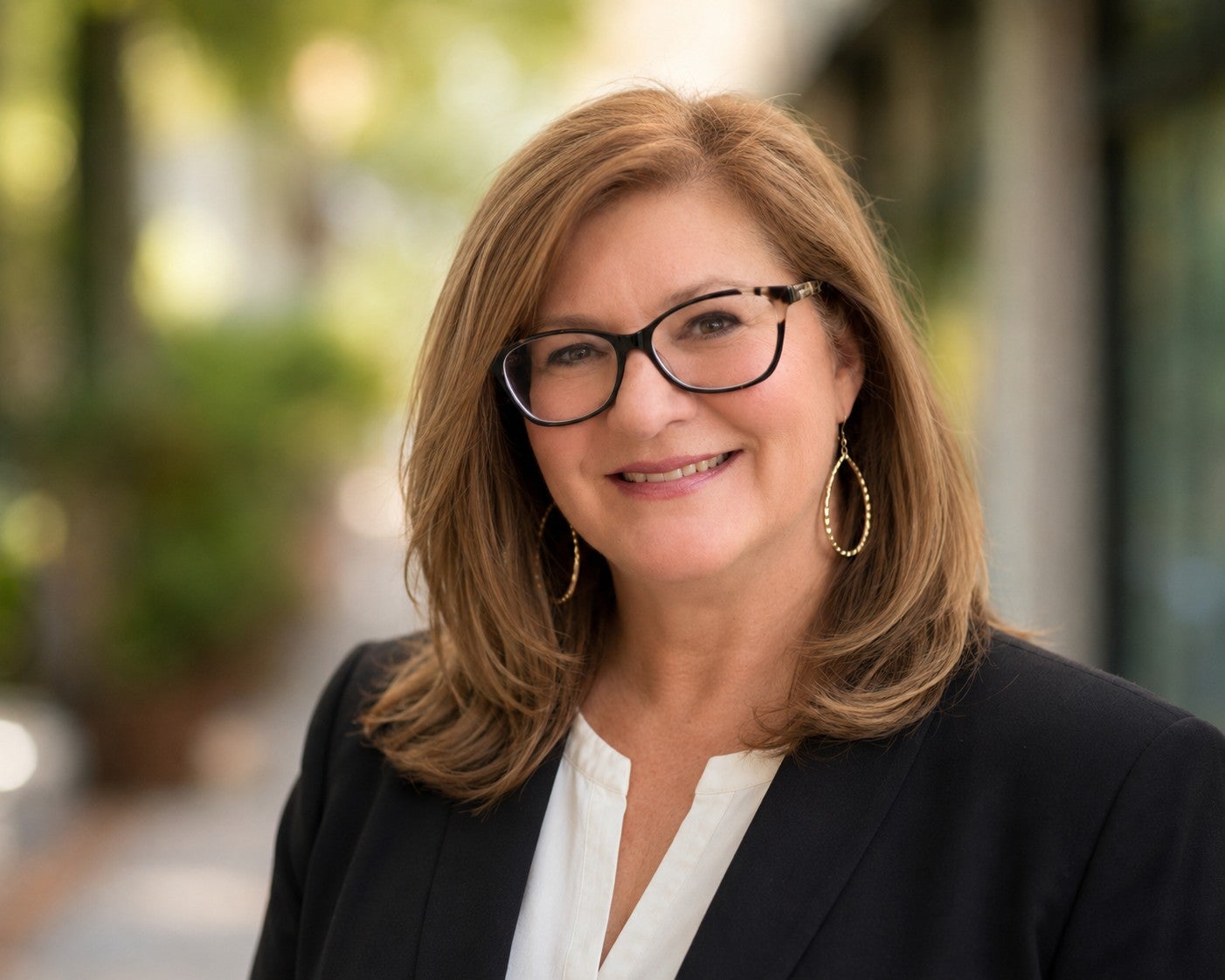 Person with sholder length, brown hair parted to the left, wearing glasses black glasses and a black and white blouse.