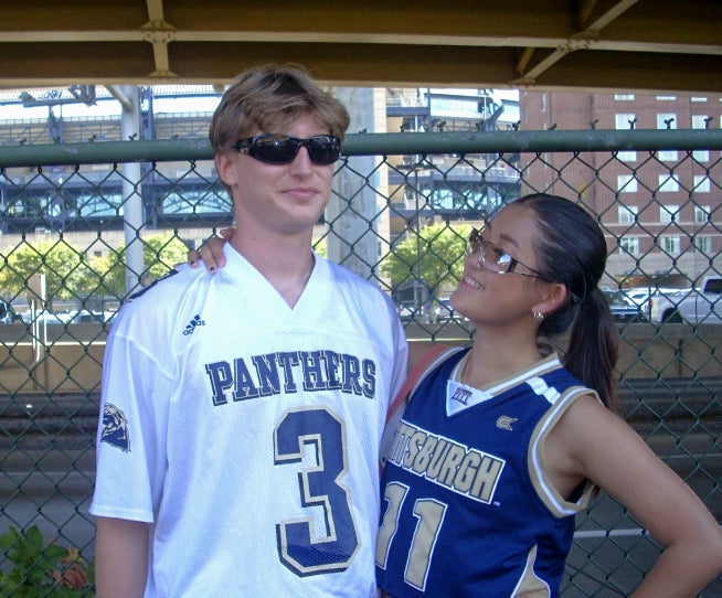 A man in a white football jersey and black sunglasses stands next to a woman in a short sleeve sports jersey wearing stylish sunglasses