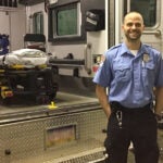 A man wearing a light blue button up shirt with a badge and American flag patch stands in front of an ambulance with its back doors open.