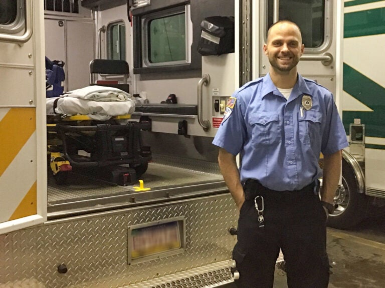 A man wearing a light blue button up shirt with a badge and American flag patch stands in front of an ambulance with its back doors open.