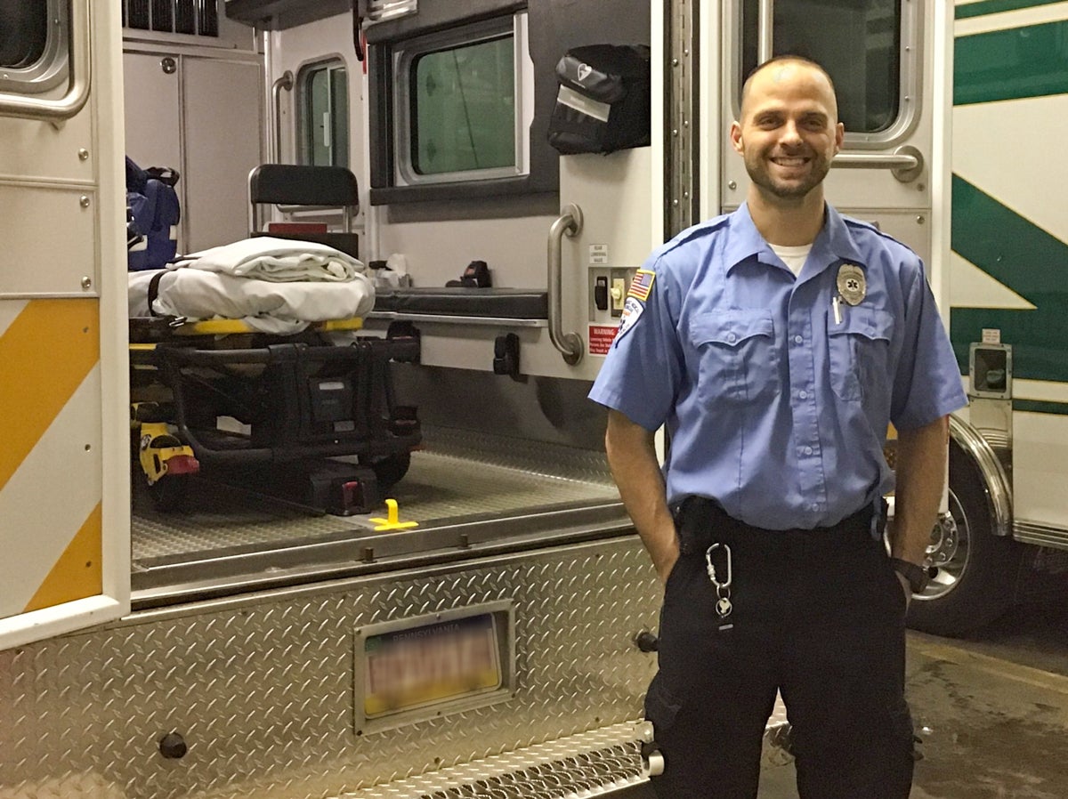 A man wearing a light blue button up shirt with a badge and American flag patch stands in front of an ambulance with its back doors open.
