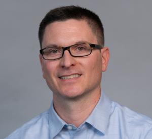Man with short dark hair wearing dark glasses and a light blue collared shirt.
