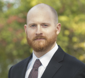 Man with very short hair and a beard wearing a black suit jacket over a white collared shirt and a brown tie.