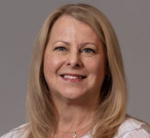 Woman with blonde hair wearing a white blouse with white and cream stripes and a silver necklace.
