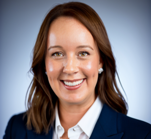 Woman with dark auburn hair wearing a black blazer over a white collared shirt and silver earrings.