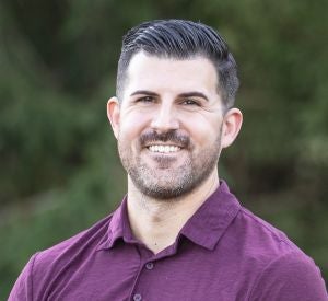 Man with dark hair and facial hair wearing a maroon collared shirt.