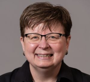 A woman with very short brown hair wearing black-rimmed glasses, a black collared blouse, silver earrings and a silver necklace.