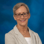 Woman with short light hair wearing dark red and brown glasses, a light tan blazer over a white blouse and a silver necklace with a pearl pendant.