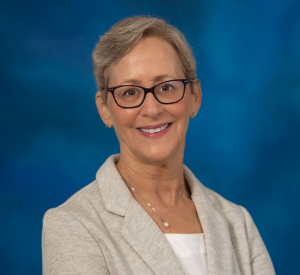 Woman with short light hair wearing dark red and brown glasses, a light tan blazer over a white blouse and a silver necklace with a pearl pendant.