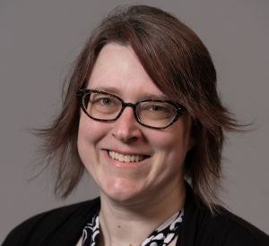 A woman with short brown hair wearing black glasses and a black cardigan over a black and white patterned blouse.