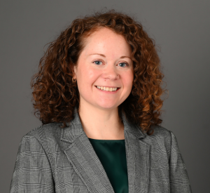 Woman with curly red hair wearing a gray blazer over a black blouse.