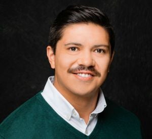 Man with dark hair and a mustache wearing a dark green swearter over a white collared shirt.