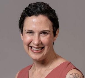 Woman with very short, dark curly hair waring a light red sleeveless shirt and a gold necklace.