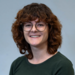 Woman with curly red hair wearing a dark green shirt and black glasses.