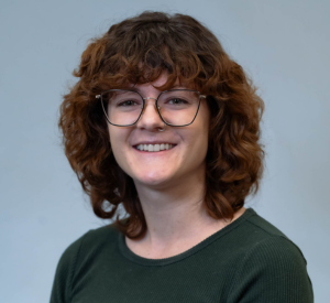 Woman with curly red hair wearing a dark green shirt and black glasses.