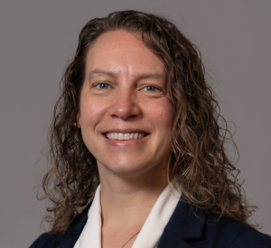 Woman with curly brown hair wearing a black blazer over a white blouse.