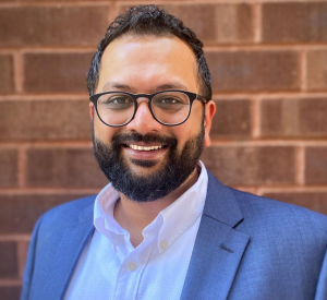 Man with dark hair and a beard wearing dark rimmed glasses and a blue-gray suit jacet over a white collared shirt.