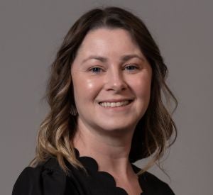 Woman with curled dark brown hair with blonde highlights wearing a black blouse with scallopped edges.
