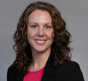 Woman with curly brown hair wearing a black blazer over a light red shirt.