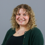 Woman with blonde curly hair wearing a green cardigan over a black shirt with a silver necklace.