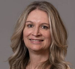Woman with light and dark blonde hair wearing a tan blouse and a gold necklace.