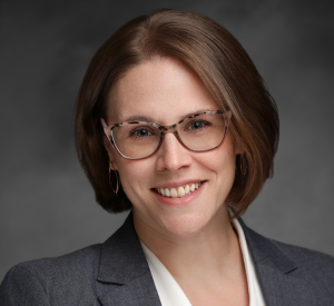 Woman with short brown hair and light brown glasses wearing a dark gray blazer over a white blouse.