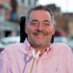 A man with short dark and gray hair and goatee wearing a light pink collared shirt