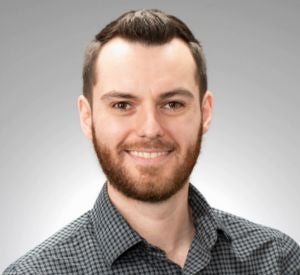 A man with short auburn hair and a beard wearing a dark grey collared shirt.