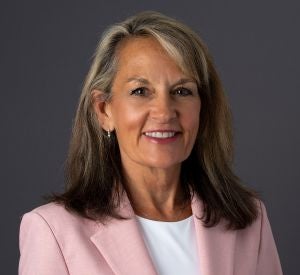 Woman with dark brown and blonde hair wearing a light pink blazer over a white shirt with silver earrings.