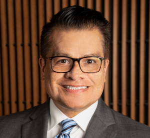 man with short dark hair wearing black glasses with a dark grey suit jacket over a white collared shirt and a dark and light blue striped tie.