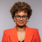 Woman with auburn curly hair wearing an orange blazer over a black shirt.
