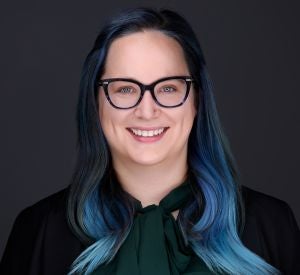 A woman with dark hair dyed blue wearing black-rimmed glasses and a black blazer over a dark green blouse.