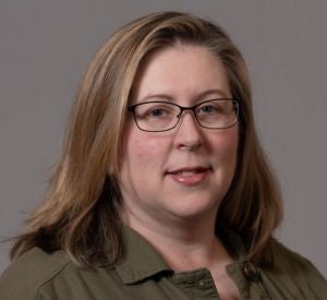 A woman with blonde hair and black wire-rimmed glasses wearing a dark green jacket over a black and white striped shirt.