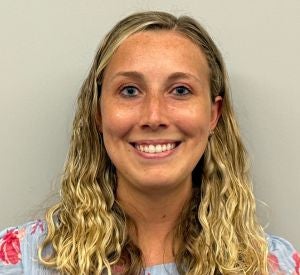 A woman with blonde curly hair pulled back wearing a light blue blouse with pink and red flowers.