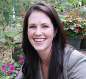 Woman with long dark hair wearing a gray shirt in front of pink and red flowers.