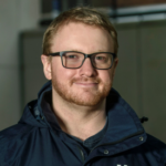 Man with reddish blonde hair and a beard wearing dark glasses and a black sweatshirt.