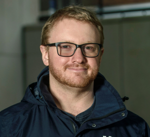 Man with reddish blonde hair and a beard wearing dark glasses and a black sweatshirt.