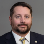 Man with short black hair and a beard wearing a black suit jacket over a white collared shirt with a gold tie with navy blue and white stripes and a University of Pittsburgh pin.